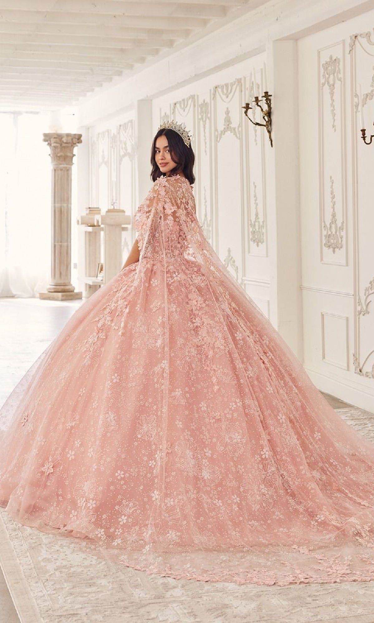 A woman stands in an ornate room wearing the Quinceanera Dress 15719 by Ladivine—a pink, floral lace ball gown with a long, lacy train and tiara—gracefully looking over her shoulder.